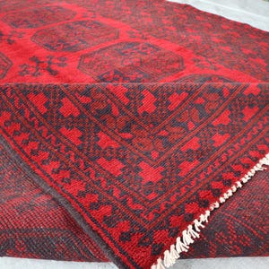 Puede incluir: Una alfombra estampada en rojo y negro con una esquina doblada, que muestra un dise&ntilde;o detallado. La alfombra presenta un patr&oacute;n geom&eacute;trico repetitivo en negro sobre un fondo rojo. La esquina doblada revela la parte inferior y el fleco de la alfombra.