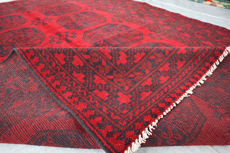 Puede incluir: Una alfombra estampada en rojo y negro con una esquina doblada, que muestra un dise&ntilde;o detallado. La alfombra presenta un patr&oacute;n geom&eacute;trico repetitivo en negro sobre un fondo rojo. La esquina doblada revela la parte inferior y el fleco de la alfombra.