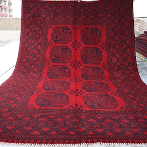 Puede incluir: Una alfombra roja rectangular grande con un patr&oacute;n geom&eacute;trico detallado en azul oscuro y negro. La alfombra presenta un dise&ntilde;o central de cuadrados conectados y un borde estampado. La alfombra se exhibe al aire libre, con una pila de alfombras similares visible en el fondo.