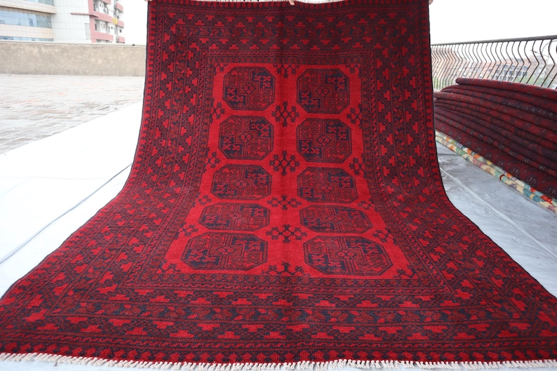 Puede incluir: Una alfombra roja rectangular grande con un patr&oacute;n geom&eacute;trico detallado en azul oscuro y negro. La alfombra presenta un dise&ntilde;o central de cuadrados conectados y un borde estampado. La alfombra se exhibe al aire libre, con una pila de alfombras similares visible en el fondo.