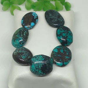 Könnte beinhalten: Eine Sammlung von acht ovalen Türkis- und braunen Edelsteinperlen, die kreisförmig angeordnet sind. Die Perlen weisen ein gesprenkeltes Muster aus Türkis und Braun auf. Die Perlen werden auf einer weißen Oberfläche mit grünem Laub im Hintergrund präsentiert.