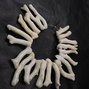 Op de afbeelding: Een streng witte koraal kralen, gerangschikt in een cirkel op een zwart lederen oppervlak. De koraal is natuurlijk en heeft een ruwe, organische textuur.