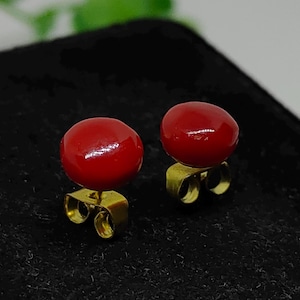 May include: A pair of red, round stud earrings. The earrings have a glossy finish and are set on gold-colored posts. The earrings are displayed on a black surface.