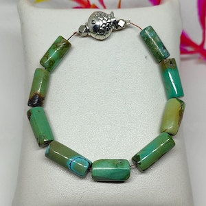 May include: A bracelet composed of cylindrical green and brown stone beads strung on a thin cord. The bracelet features a silver-toned metal clasp shaped like a fish, with detailed scale patterns and a small eye visible on the surface of the metal.
