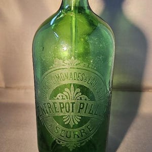 Puede incluir: Una botella de sifón de soda de vidrio verde vintage con un cabezal dispensador de tono plateado metálico. El cuerpo de vidrio presenta un emblema circular grabado con el texto BIERES LIMONADES & EAUX DE SELZ, ENTREPOT PILLON, A SEURRE.