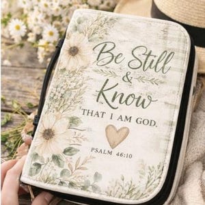 Pode incluir: Uma capa de Bíblia creme com um design floral e o texto "Be Still & Know THAT I AM GOD." A capa tem um zíper e uma alça pretos. Salmo 46:10 também está impresso na capa.