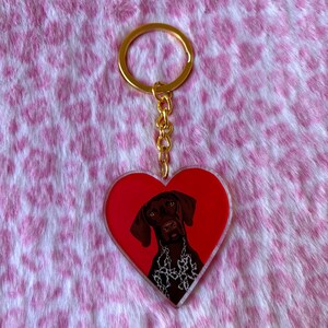 May include: A heart-shaped keyring with a brown dog portrait on a red background. The dog has a white and black patterned collar. The keyring has a gold-coloured ring and chain.