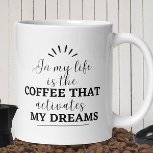以下が含まれることがあります： 白いセラミック製のコーヒーカップに、黒い文字で「In my life is the COFFEE THAT activates MY DREAMS」と書かれています。カップはコーヒー豆の上に置かれています。
