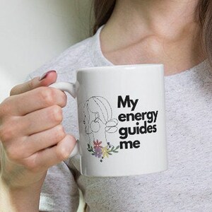 Könnte beinhalten: Weiße Keramiktasse mit einer schwarzen Umrandung eines Frauenkopfes und dem Text "My energy guides me" mit einem floralen Design darunter.