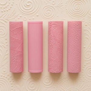May include: Four pink rolling pins with textured designs. The patterns include leaf and dotted textures, and vertical lines. The rolling pins are cylindrical and stand upright against a cream-coloured background with embossed floral patterns.