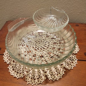 May include: A two-tiered glass serving dish shaped like a seashell. The top tier is smaller and sits inside the larger bottom tier. Both tiers are clear glass and have a rippled texture.
