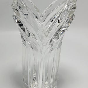 May include: Clear glass vase with a unique, angular design. The vase has a square base and a flared, V-shaped top. Vertical ridges run along the sides, adding texture. The overall height is approximately 10 inches.