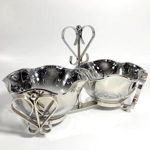 May include: A silver-toned serving dish with two scalloped bowls. The bowls are connected by a decorative handle with heart-shaped accents. The dish has a reflective surface and is designed for serving condiments or small portions.