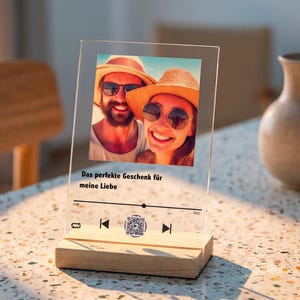 Könnte beinhalten: Ein klarer Acryl-Fotorahmen mit Holzsockel. Er zeigt ein Farbfoto eines lächelnden Paares mit Sonnenbrille und Hut. Der Text "Das perfekte Geschenk für meine Liebe" ist darunter gedruckt, zusammen mit einem QR-Code.