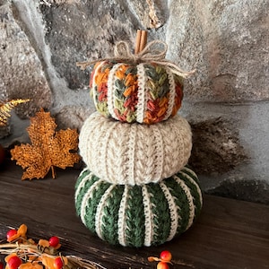 May include: A stack of three crocheted pumpkins featuring a textured cable stitch pattern. The top pumpkin is multicolored with orange, green, and yellow yarn, the middle is cream, and the bottom is dark green. A cinnamon stick stem sits on top.