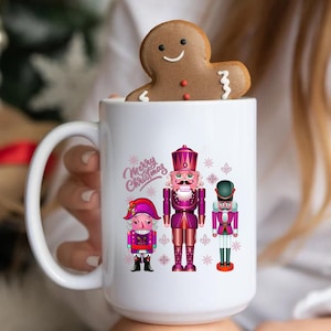 Könnte beinhalten: Weiße Keramiktasse mit einem rosa-weißen Design mit drei Nussknackerfiguren und dem Text "Frohe Weihnachten".