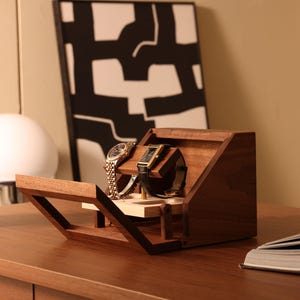 May include: A dark brown wooden watch display stand with an angled front panel and a horizontal bar holding two wristwatches. The stand features a light-colored wooden base platform and a hinged front frame that extends outward from the main body.