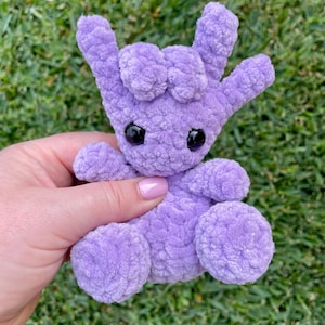 May include: A lavender-coloured crocheted plush toy in the shape of a hand making the "I love you" sign. The toy has black safety eyes and is made of soft, textured yarn. The background is green grass.