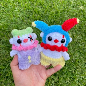 May include: Two crocheted plush toys shaped like clowns. One is wearing a blue and red jester hat and yellow pants. The other is wearing a pink and purple jester hat and purple pants.