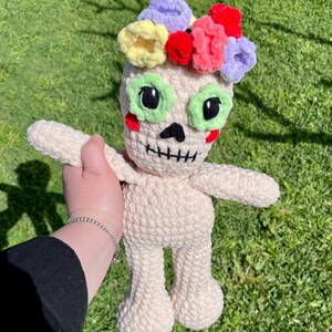 May include: A crocheted doll with a white body and a skull face. The doll is wearing a colorful flower crown with pink, yellow, green, and purple flowers.