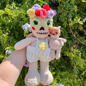 May include: A crocheted plush toy in the shape of a skeleton wearing a floral headband and overalls. The toy is holding a smaller crocheted plush toy in the shape of a cat.