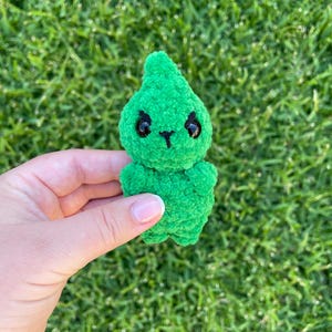 Könnte beinhalten: Ein kleines, handgemachtes grünes Häkeltier mit schwarzen Augen und einem spitzen Kopf. Das Plüschtier wird vor einem Hintergrund aus grünem Gras gehalten.