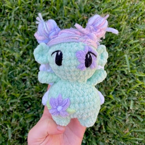 Puede incluir: Un juguete de peluche tejido a crochet con un cuerpo verde, flores moradas y pelo de lana rosa y blanco. El juguete tiene grandes ojos negros y una expresión amigable.