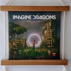 Op de afbeelding: Een houten platenspelerdisplay met een wit touw dat van boven hangt. Het display bevat een vinylplaat met een afbeelding van een man die in een veld loopt met een gebouw op de achtergrond. De platenhoes zegt "Imagine Dragons Origins".