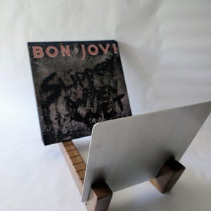May include: A record stand made of wood and metal, holding a Bon Jovi album. The album cover is black and grey with pink text. The stand has a rectangular wooden base and a silver metal back.