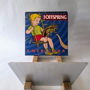 May include: A vinyl record album cover for "Americana" by The Offspring. The artwork features a cartoon boy on a swing with a large insect. The album title and band name are displayed in red and white text against a blue background.