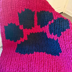 Può includere: Una coperta lavorata a mano a maglia grossa, di colore fucsia vibrante, con un grande motivo a impronta di zampa blu navy. La coperta è realizzata in filato morbido e spesso, perfetto per coccolarsi. Il motivo a impronta aggiunge un tocco giocoso.