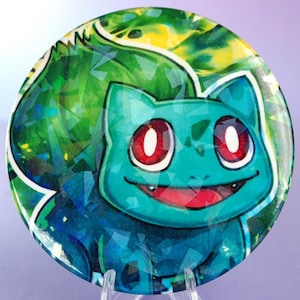 Peut inclure: Une assiette décorative ronde avec un design de personnage Bulbasaur. L'assiette présente un Bulbasaur turquoise avec des yeux rouges et une expression souriante, sur un fond vert et jaune. L'assiette est présentée sur un présentoir en acrylique transparent.