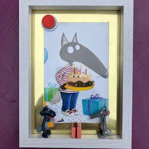 May include: Framed artwork with a cartoon wolf holding a birthday cake. The image includes a red button, small gift boxes, and miniature wolf figurines. The white frame has a gold border, set against a purple background.