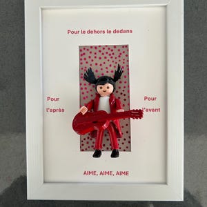 Op de afbeelding: Een ingelijst kunstwerk met een Playmobil-figuur in een rood pak met een rode gitaar. De figuur staat tegen een roze achtergrond met stippen. Franse tekst omringt de figuur, met de woorden "AIME, AIME, AIME" onderaan.
