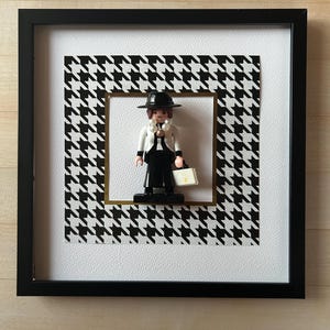 Peut inclure: Une image encadrée en noir et blanc avec une bordure en motif pied-de-poule noir et blanc. L'image présente une figurine en noir et blanc d'une personne portant un chapeau noir, une chemise blanche et un pantalon noir. La figurine tient une mallette blanche.