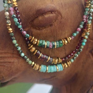 Peut inclure: Trois colliers superposés avec des perles de pierres précieuses dans les tons de vert, de rouge et de turquoise, rehaussés de perles en métal doré. Les colliers sont présentés sur un fond en bois.
