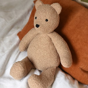 Puede incluir: Un oso de peluche marr&oacute;n claro con una superficie texturizada, ojos y nariz negros. El peluche est&aacute; sobre una colcha blanca, con un coj&iacute;n de pana color &oacute;xido detr&aacute;s. Un juguete para ni&ntilde;os.