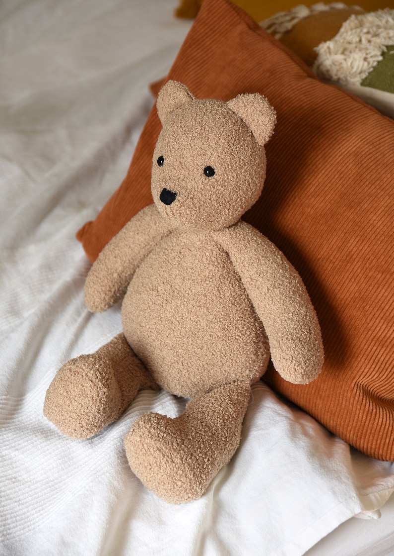 Puede incluir: Un oso de peluche marr&oacute;n claro con una superficie texturizada, ojos y nariz negros. El peluche est&aacute; sobre una colcha blanca, con un coj&iacute;n de pana color &oacute;xido detr&aacute;s. Un juguete para ni&ntilde;os.
