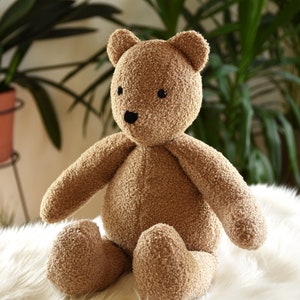 Puede incluir: Un oso de peluche marr&oacute;n con un exterior texturizado y difuso. El oso tiene peque&ntilde;os ojos negros y una nariz negra. Est&aacute; sentado sobre una superficie blanca y esponjosa, con plantas verdes en el fondo.