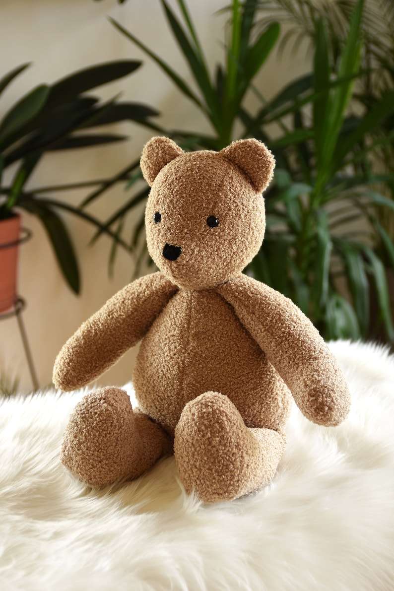 Puede incluir: Un oso de peluche marr&oacute;n con un exterior texturizado y difuso. El oso tiene peque&ntilde;os ojos negros y una nariz negra. Est&aacute; sentado sobre una superficie blanca y esponjosa, con plantas verdes en el fondo.
