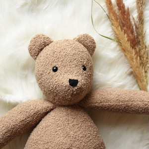 Puede incluir: Un oso de peluche hecho a mano con una tela suave y texturizada de color marr&oacute;n claro. El oso tiene peque&ntilde;os ojos negros y una nariz negra. Los brazos del oso est&aacute;n extendidos y est&aacute; acostado sobre una superficie blanca y esponjosa. Hay hierbas secas en el fondo.