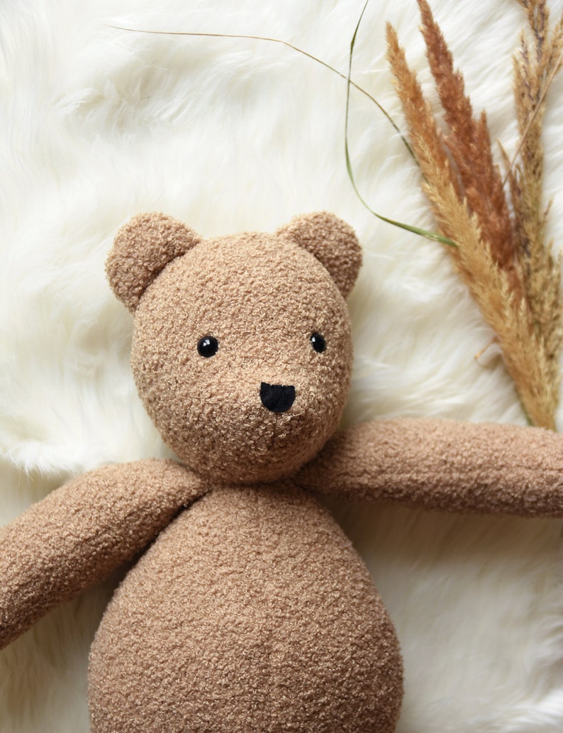 Puede incluir: Un oso de peluche hecho a mano con una tela suave y texturizada de color marr&oacute;n claro. El oso tiene peque&ntilde;os ojos negros y una nariz negra. Los brazos del oso est&aacute;n extendidos y est&aacute; acostado sobre una superficie blanca y esponjosa. Hay hierbas secas en el fondo.