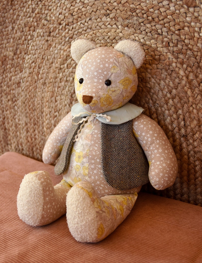 Puede incluir: Un oso de peluche hecho a mano con un cuerpo estampado floral y un chaleco de tweed. El oso tiene un cuello azul claro y patas y orejas texturizadas de color crema. El oso est&aacute; sentado sobre una superficie rosa.