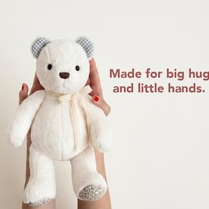 May include: A cream-coloured plush teddy bear with dark eyes and a brown nose. The bear features blue and white gingham inner ears, floral patterned foot pads, and a cream ribbon tied around its neck. The text reads: Made for big hugs and little hands.