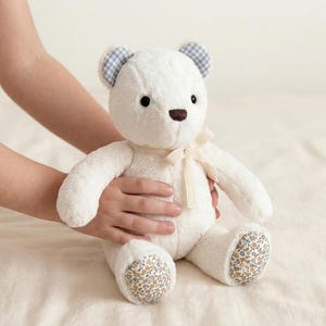 May include: A plush teddy bear with white fur, featuring blue and white gingham patterned inner ears, a brown nose, and a cream-coloured ribbon tied around its neck. The bear has floral patterned fabric on the soles of its feet with small flowers.