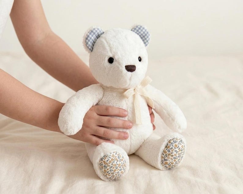 May include: A plush teddy bear with white fur, featuring blue and white gingham patterned inner ears, a brown nose, and a cream-coloured ribbon tied around its neck. The bear has floral patterned fabric on the soles of its feet with small flowers.