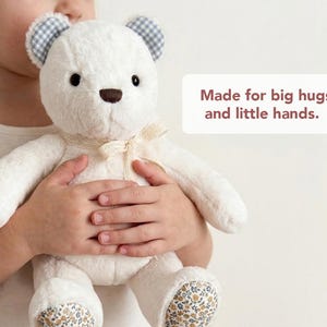 May include: A plush white teddy bear with a brown nose, blue and white checked ears, and floral patterned feet. A cream-coloured lace ribbon is tied around its neck. The text "Made for big hugs and little hands." is printed on the right side.