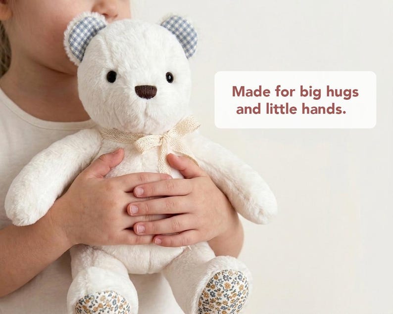 May include: A plush white teddy bear with a brown nose, blue and white checked ears, and floral patterned feet. A cream-coloured lace ribbon is tied around its neck. The text "Made for big hugs and little hands." is printed on the right side.