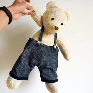 Puede incluir: Un oso de peluche hecho a mano con un cuerpo estampado floral y un peto vaquero. El oso tiene un fondo beige claro con acentos florales amarillos, ojos de bot&oacute;n negros y una nariz marr&oacute;n. El peto vaquero tiene cierres de bot&oacute;n de madera.