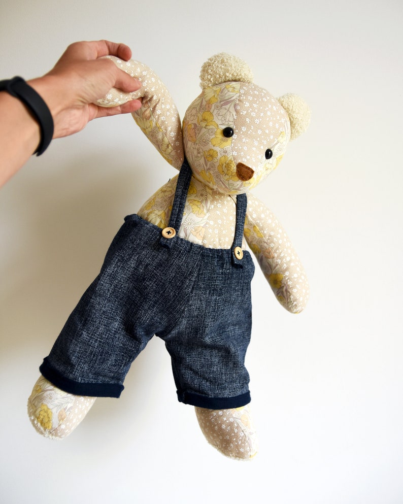 Puede incluir: Un oso de peluche hecho a mano con un cuerpo estampado floral y un peto vaquero. El oso tiene un fondo beige claro con acentos florales amarillos, ojos de bot&oacute;n negros y una nariz marr&oacute;n. El peto vaquero tiene cierres de bot&oacute;n de madera.